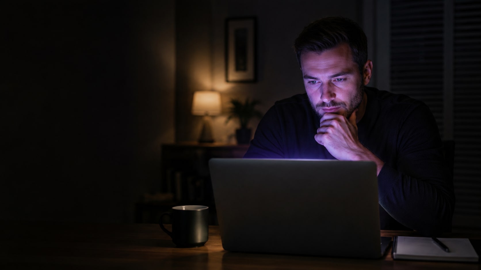 A person at a home desk late at night, seen from behind their laptop so only the lid faces us, the screen's soft glow lighting a thoughtful face in a dark room.