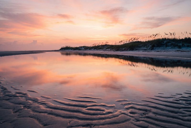 A mirror-flat tidal pool at first light reflecting a pink dawn sky, sand ripples leading into it.