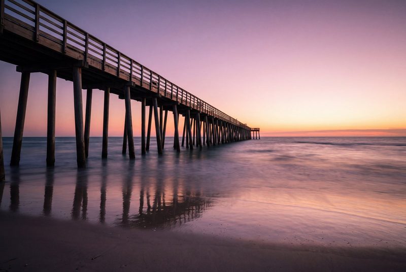 A fishing pier running out over silk-smooth water at blue hour, its pilings reflected in the wet sand.